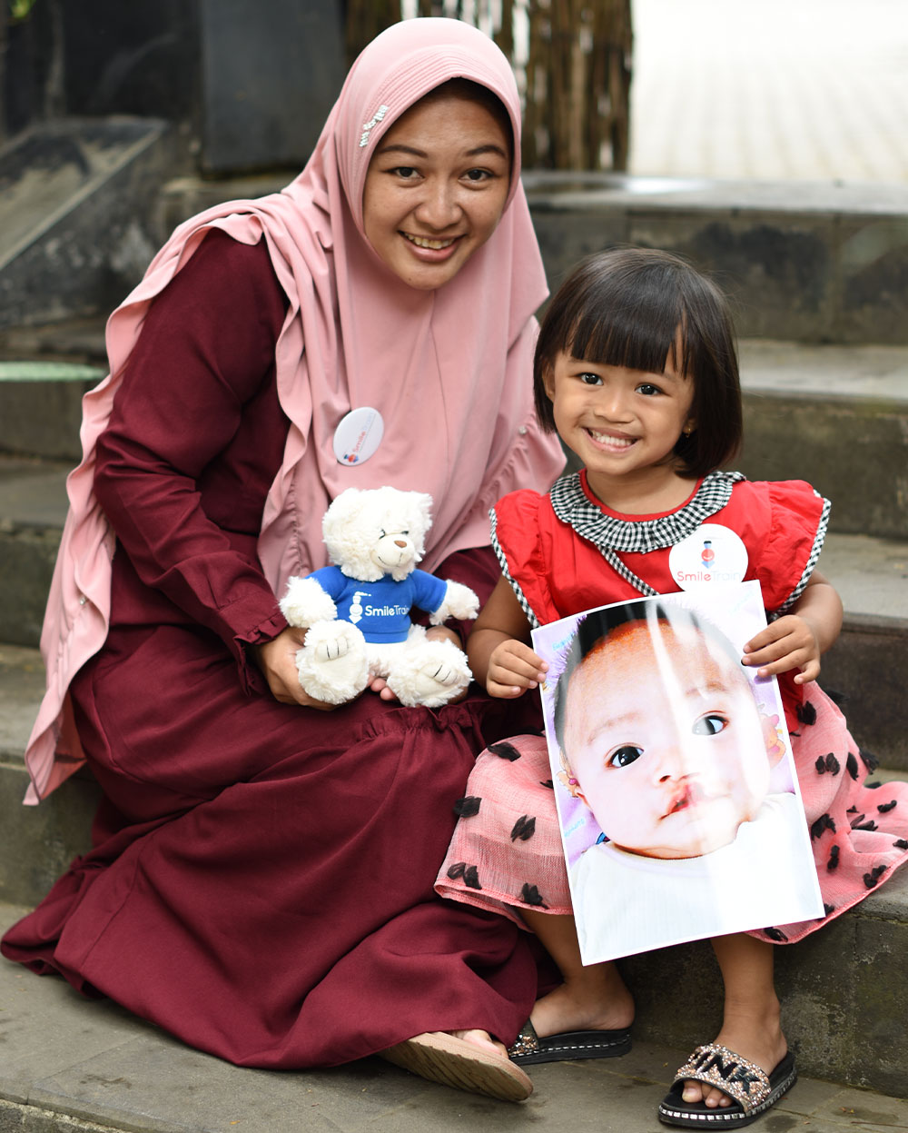 Ruri and Khalisa smiling and holding a teddy bear and a picture of Khalisa before cleft surgery